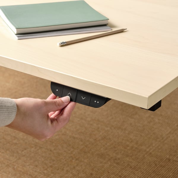 Hand pressing down button on black control panel under beige desk, adjusting height for sit/stand.