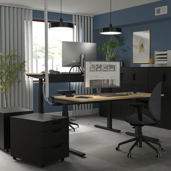 Modern home office: MITTZON black sit-stand desk, grey chair, dual monitors, blue walls, potted plant.