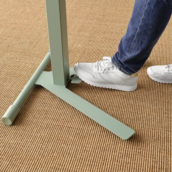 A persons feet, wearing jeans and white trainers, stand on a green, height-adjustable laptop table with a tiltable top.