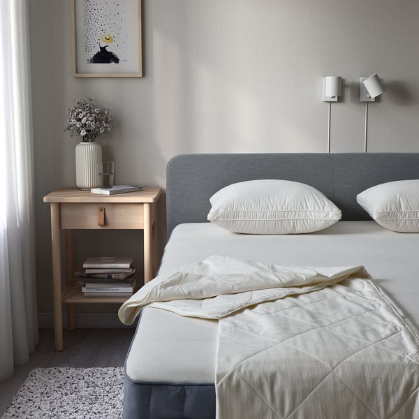 Twin bed, grey headboard, quilt, 2 pillows. Bedside table with flowers, candle, books. 2 wall lamps, framed pic, sheer curtains. Simple, serene.