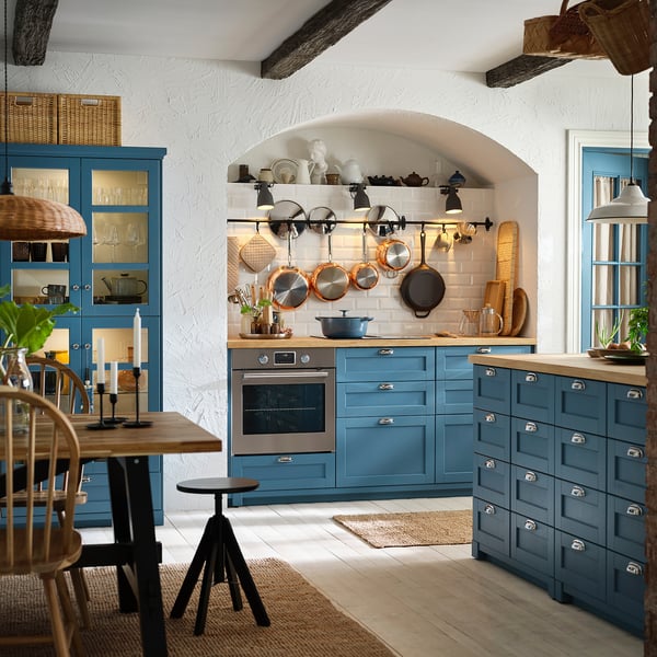 Modern kitchen with blue cabinets, oak countertop, hanging pots, wooden dining table, and wicker chairs.