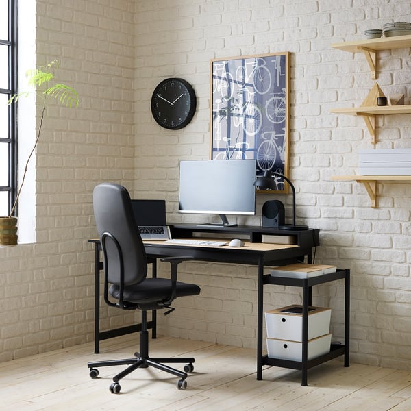 Black modern office setup with grey chair & monitor. White brick wall backdrop. Wooden shelves hold books & decor.