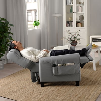 A person reclines in a comfortable MUREN armchair with adjustable back and footrest extended, set in a cosy living room with bookshelves and a window.