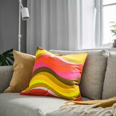 Brightly coloured wave-patterned throw pillow on grey couch, near large window. Brown and neutral pillows nearby.