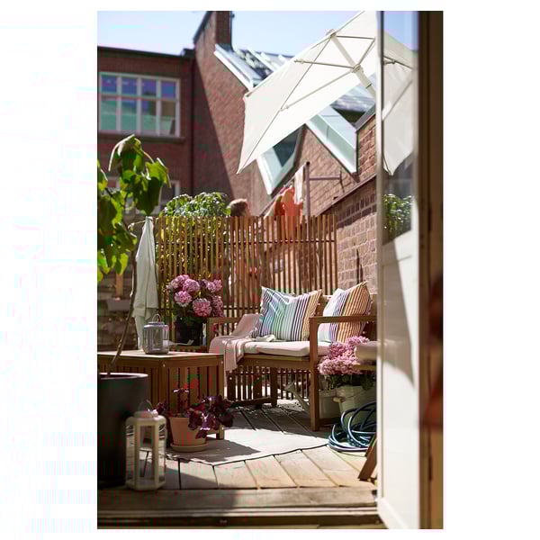 Wooden patio set on balcony with cushions, white flowers, bench & table under an umbrella, brick wall & plant.
