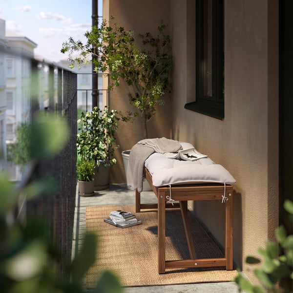 Small outdoor balcony with brown NÄMMARÖ bench, white cushion, green plants, beige rug, and black railing.