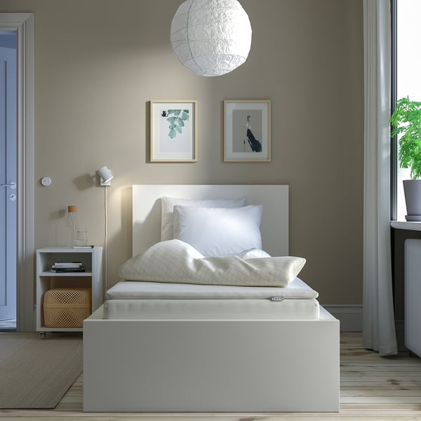 Modern bedroom with white bed on grey frame, bedside table, lamp, books, and plant.