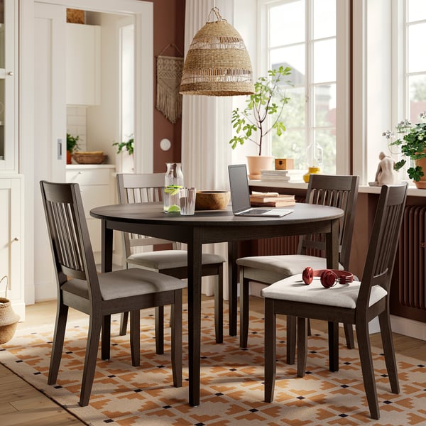 Modern farmhouse dining set with dark grey round table and 4 cushioned chairs, placed on a multicoloured rug under a large window.
