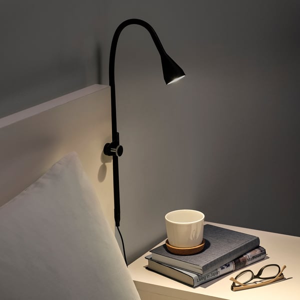 Black adjustable clamp lamp on bedside, illuminating a stack of books with a mug on top.