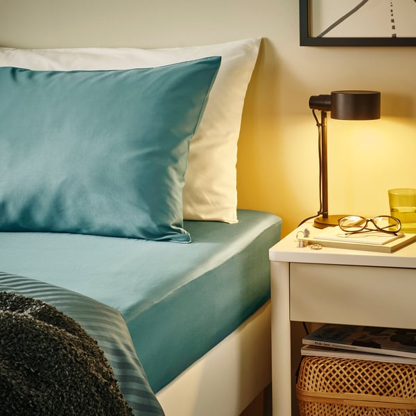 Teal bed, white pillows, cosy blanket. Beside it, a bedside table with lamp, glasses, and a cup.