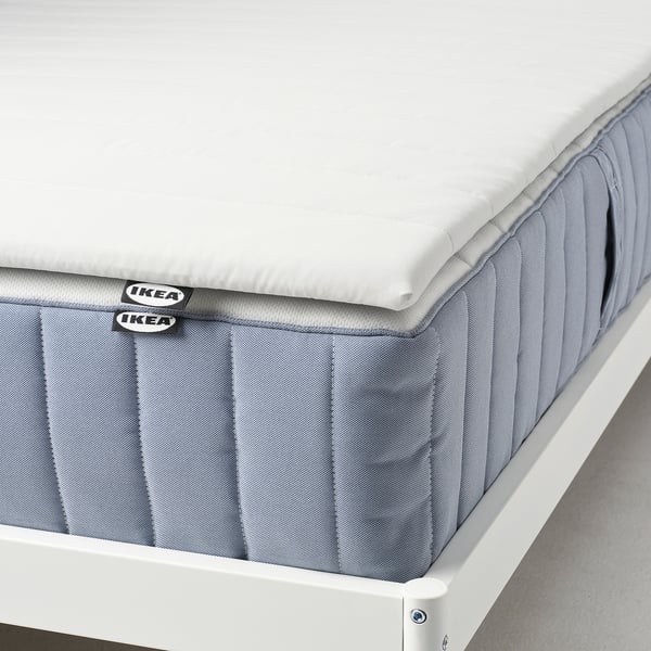 Displays a rolled mattress pad with a grey cover on a white bed frame, showing its comfort and protective.