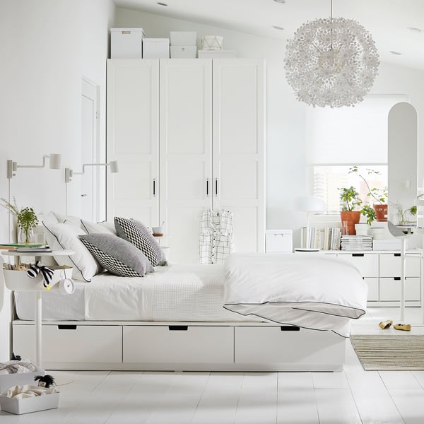 Modern minimalist white bedroom, featuring NORDLI bed with storage, pendant lamp, black-and-white pillows, side tables with plants, and large mirror.