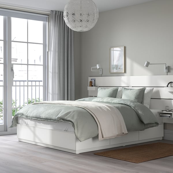 Modern minimalist bedroom with white NORDLI bed, grey bedding, spherical chandelier, headboard shelves, and large window.