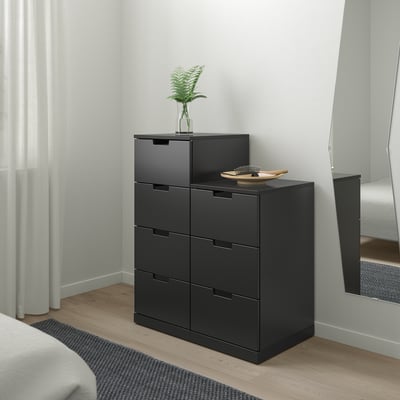 Black NORDLI chest with six drawers, plant, bowl. Silent runners. Beside bed, white wall.