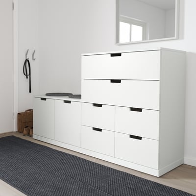 Modern white NORDLI chest in hallway, with grey rug. Tall, six-drawer unit with black handles, placed near hooks and bench.