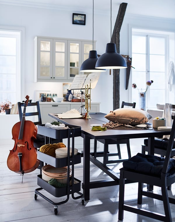 Bright dining room with NORDVIKEN black chairs, wooden table, hanging lights, musical instruments, and kitchen in background.