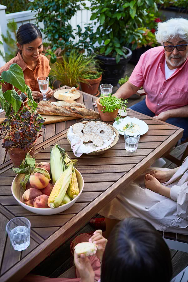 Family enjoying meal outdoors on wooden table with bread, fruits, and glasses.