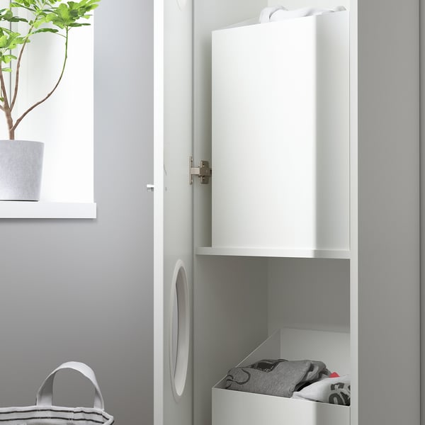 White cabinet for laundry, with open door showing folded clothes in bottom bin, next to potted plant and shelf.