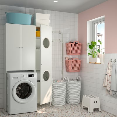 Modern bathroom with white washing machine, drying racks, and pink-painted walls. Storage cabinets, laundry baskets, and a small plant.