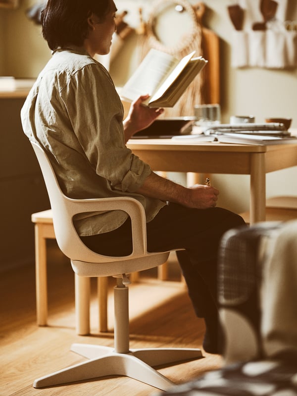 Person sitting on ODGER white swivel office chair, reading a book at a desk, wearing a light green shirt and dark pants.