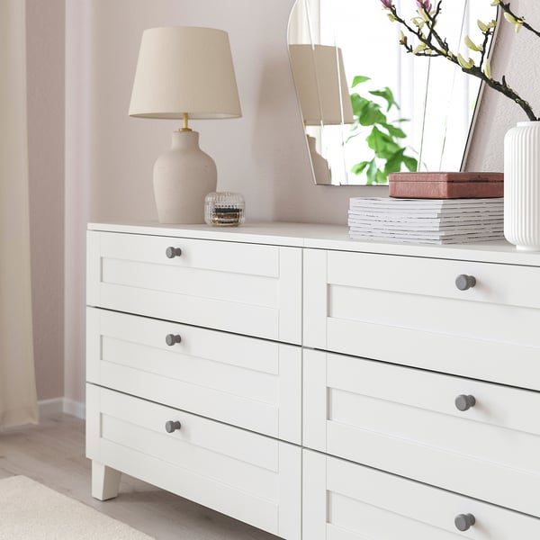 White dresser with six drawers and grey knobs, topped with a lamp, books, and a vase.