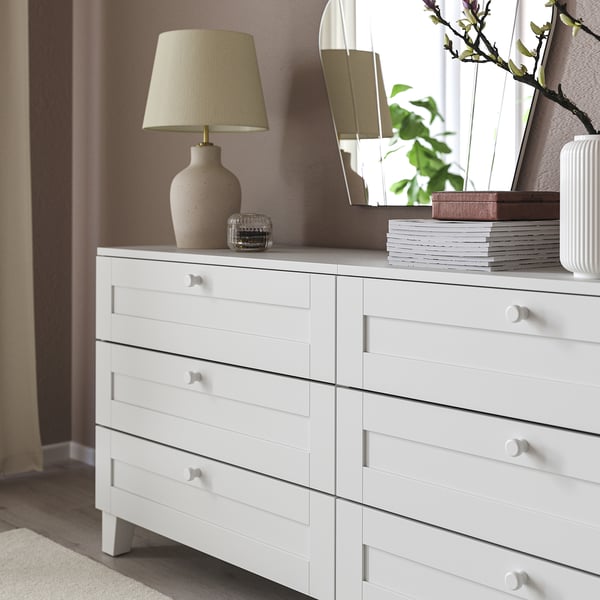 White dresser with six drawers and round knobs, topped with a lamp, decorative vase, candle, stack of books, and mirror.