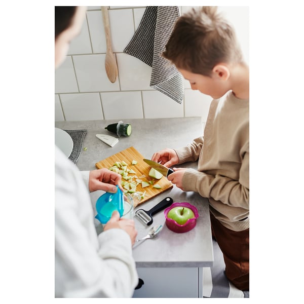 Two people chop apples on a counter. One uses a blue silicone cover on glass. Counter has peeler, whole apple, and cloth. White-tiled background.
