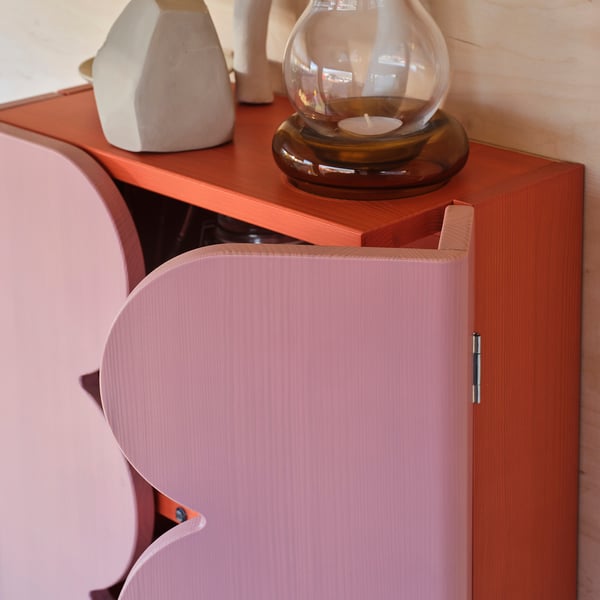 A wooden cabinet in pink and orange tones, featuring an open door, with a decorative candle and vases on top.