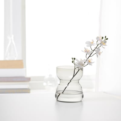 Small glass vase with bulbous base, holding white flowers on white table, with books and decor in background.