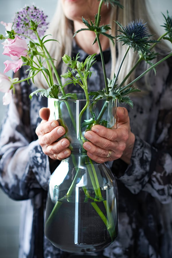 Person holding clear glass vase with long stemmed flowers, including purple and pink blossoms.