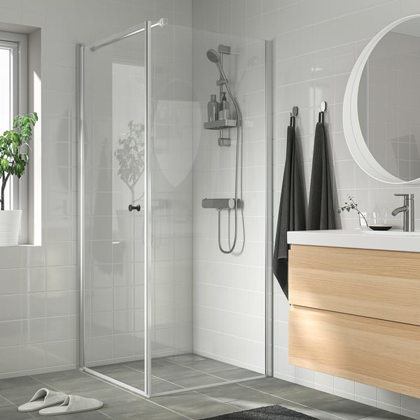 Modern minimalist bathroom with glass shower, slim silver frame, wooden vanity, mirror, black towels, plant, dark grey tiles.