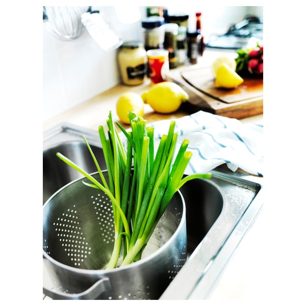Image a stainless steel sink with green onions in a colander, lemons, and kitchen items on the countertop.