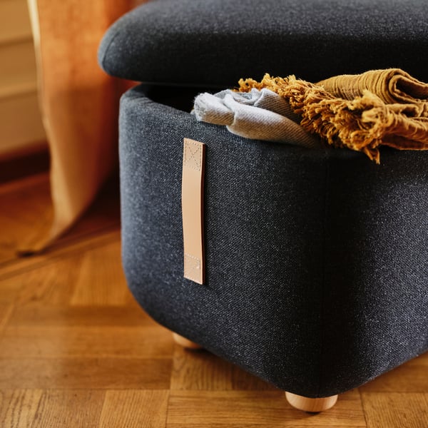 Home storage: dark grey cushioned footstool, half open revealing folded blankets. Beside orange chair on wooden floor.
