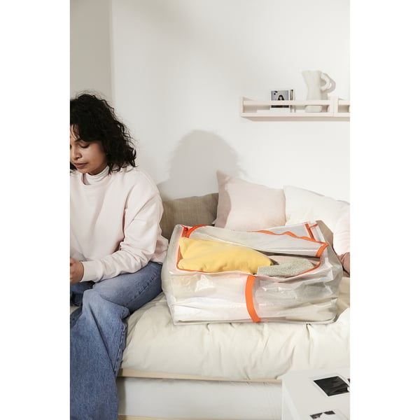 Person sitting on bed with large PÄRKLA storage case, containing folded yellow item and grey folds. White wall and shelf in background.