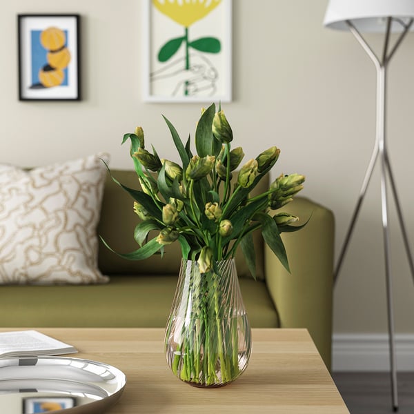 Glass PÅSKÖTRÄD vase with swirls holding yellow-green flowers on wooden table next to lamp and couch.