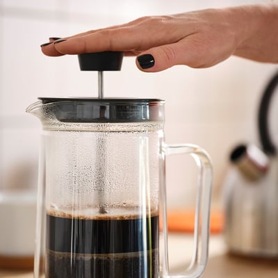 A person presses a plunger in a clear cafetière coffee maker. Dark coffee is visible inside.