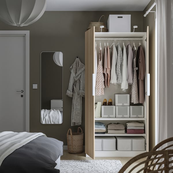 Open wardrobe displaying clothes, storage boxes, and a robe. Adjacent mirror and door evident, with a bed and chair partially seen.