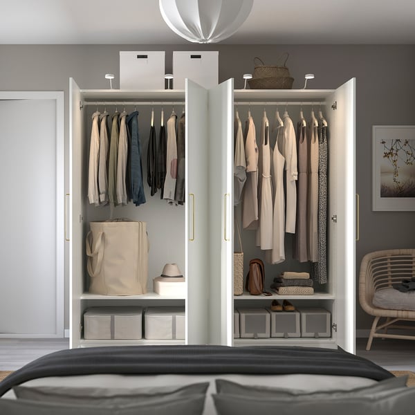 Well-organized white wardrobe with clothes, bags, and bins, placed beside a bed. Hinged doors, ample storage, and shelves visible.