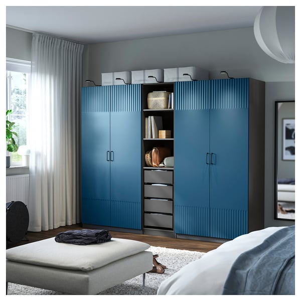 A room with a tall, blue PAX wardrobe, larger than the white ottoman in front. It has textured doors and open shelves showcasing various items.