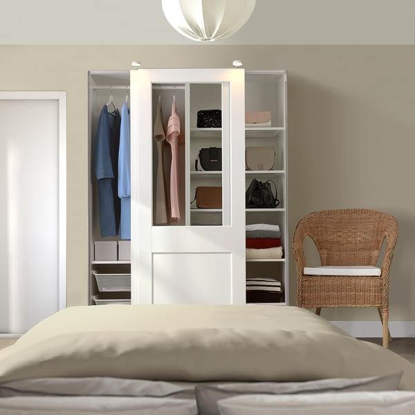 Bedroom with white PAX wardrobe and glass doors, organised clothes, beige bed, wicker chair.