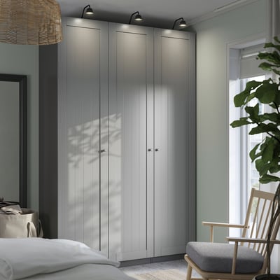Modern bedroom with tall grey PAX wardrobes, lit tops, white bed, wooden chair, and large plant by the window.
