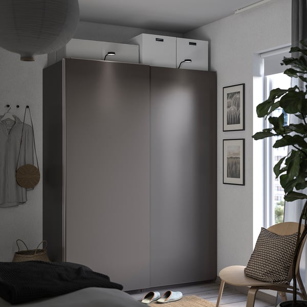 A modern bedroom with dark grey PAX wardrobe, white boxes, a wooden chair, a plant, and framed photos. A cosy and organised space.