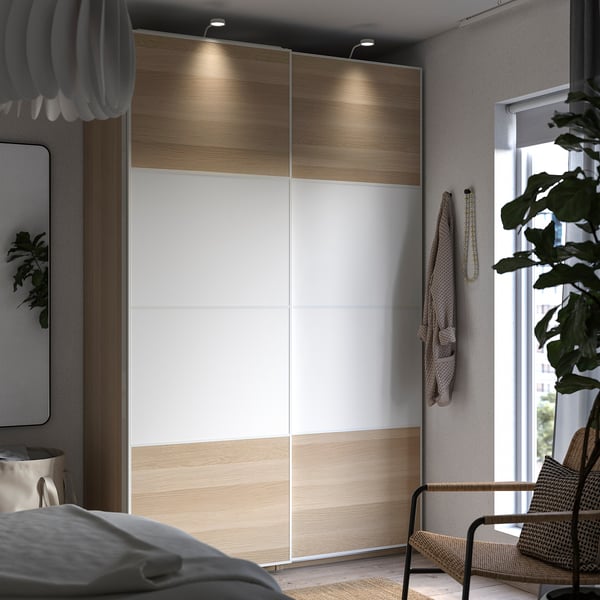 Modern bedroom with white and wooden PAX wardrobe, sliding doors, grey bedding, wicker chair, and houseplant by the window.