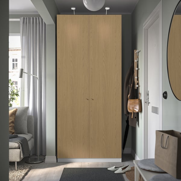Modern light brown PAX wardrobe near white door, taller than grey chair and lamp.