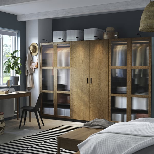 Bedroom with large PAX wardrobe, glass doors, black desk, plant. Wardrobe topped with storage, bed with white blanket.