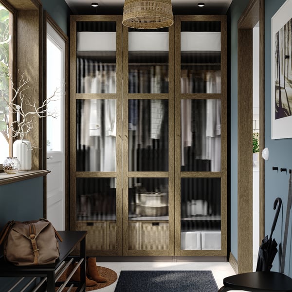 Large wooden closet, glass doors, modern design. Sits between windows, near entrance. Dark tones, tall, wide, with top and bottom shelves.