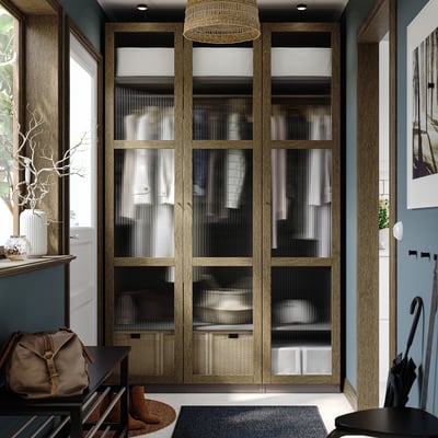 Large wooden closet, glass doors, modern design. Sits between windows, near entrance. Dark tones, tall, wide, with top and bottom shelves.