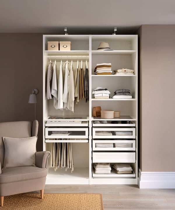 Modern walk-in closet with white PAX shelves. Hangs clothes, stacks folded items, stores accessories. Wood floor, chair left.