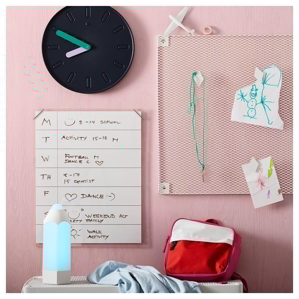 Organised kids area: wall clock, whiteboard, bulletin board with drawings, red backpack, folded blanket, blue lamp.