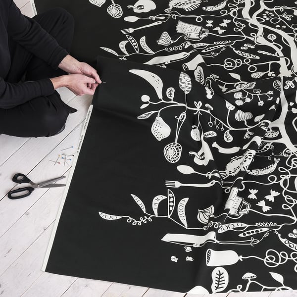 Person holds black fabric with white playful patterns, scissors and pins laid out, suggesting preparation for a sewing project.